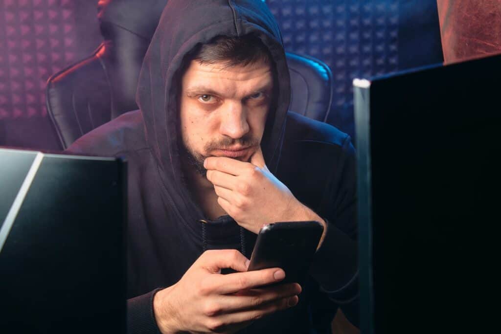 Man in a black hoodie looks at a cell phone between two desktop computers. Cyber Security vs Computer Science