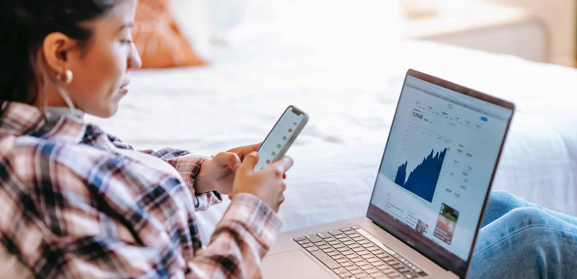 Woman sits with a laptop and uses a smartphone. How to Become an Operations Analyst: A Step-by-Step Guide