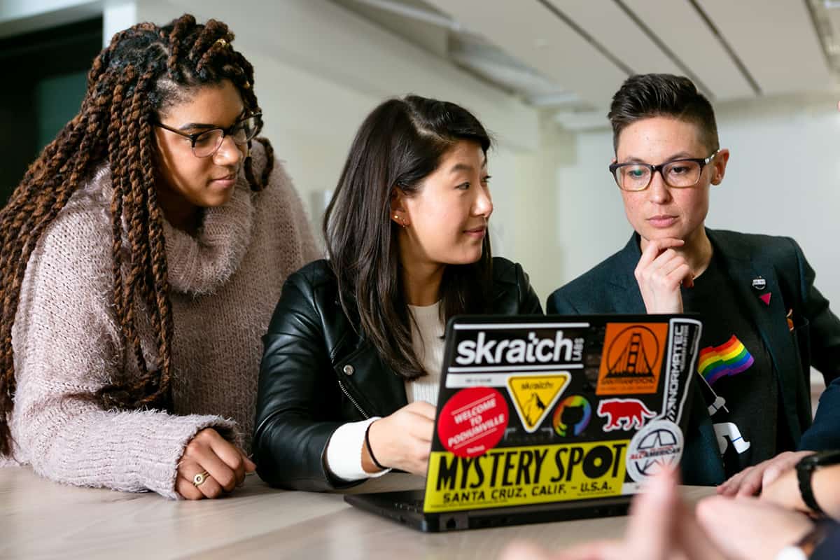 Three students in a discussion in front of a laptop. LGBTQ+ Coding Bootcamp Scholarships