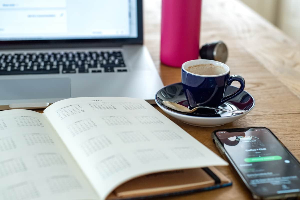 A work table with a calendar, a smartphone, a teacup and saucer, and a laptop.